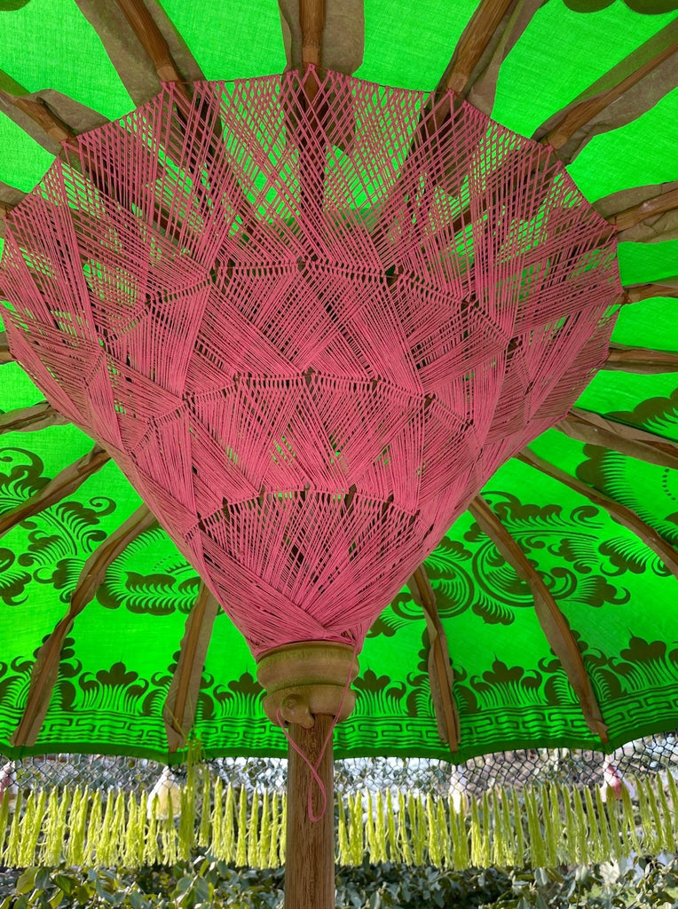 The Samantha Parasol by Baliaric Parasol exemplifies tropical elegance with its lively green canopy adorned with detailed pink string patterns. Supported by wooden spokes, this artisan parasol features decorative floral designs, providing a chic and stylish shade on a sunny day.