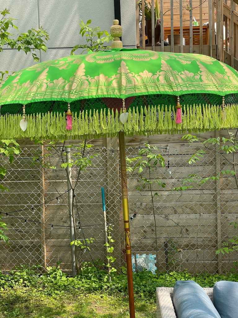 The Samantha Parasol by Baliaric Parasol, featuring exquisite gold patterns and tassels, stands open in a garden, exuding tropical elegance. Behind it is a wooden fence draped with lush greenery. The ground is carpeted with green grass, and a hint of a blue chair peeks into view, adding a splash of color to the serene scene.