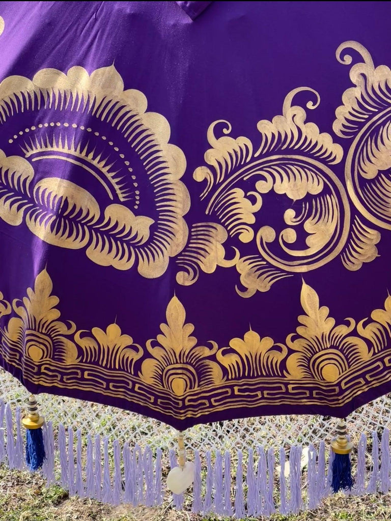 A close-up of the Claudia Parasol by Baliaric Parasol showcases its ornate purple fabric adorned with intricate gold patterns and tassels, capturing the elegance of a handcrafted Balinese parasol. The design features fans and floral motifs, creating an elaborate look that is perfect for luxurious outdoor decor.