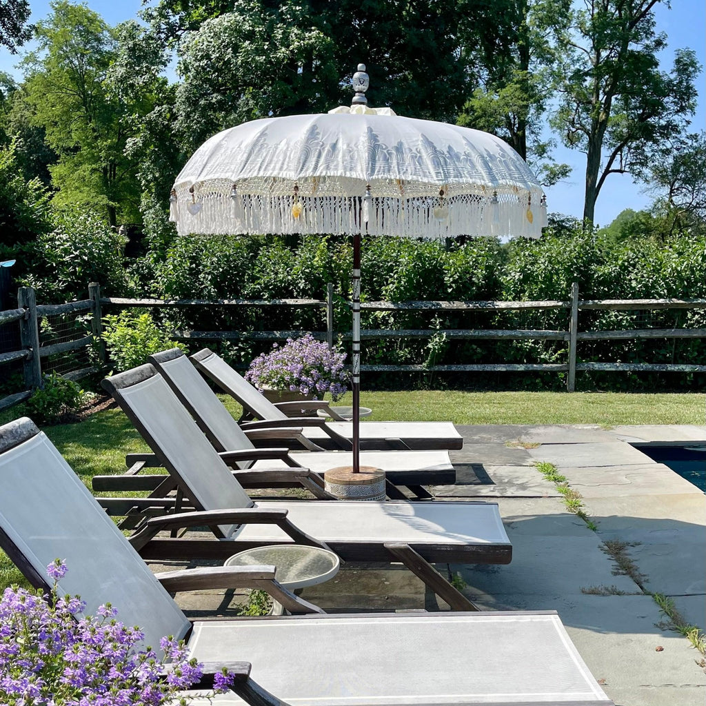 A serene poolside scene showcases five empty lounge chairs beneath a Baliaric Parasol's White Cloud Parasol on a stone patio. Vibrant color is added by purple flowers, while trees create a lush backdrop, crafting the perfect outdoor living space to unwind.
